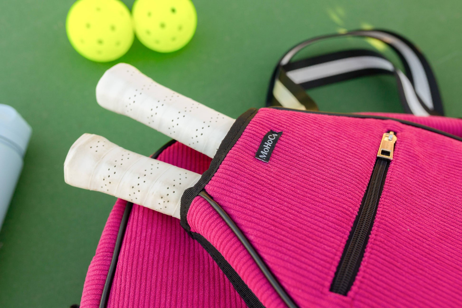 Pink sports bag with tennis rackets, squash balls, and a water bottle on a green surface
